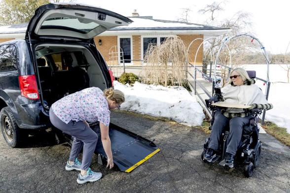 Photo taken outside on a snowy day of a woman about to enter a car with the assistance of a support worker, who is setting up a wheelchair ramp.
