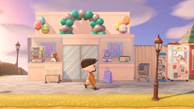 Animal Crossing: New Horizons screenshot. Image shows a bald, light skinned, and bearded human seen in a brown jacket walking from the side view. He is walking past a faux building made from wide display stands (white). In front are two windows with custom signs that read "Toy Store" in colorful letters. In the windows are assorted toys. Immediately to the right of the building is a shopping cart and toy based vending machines.