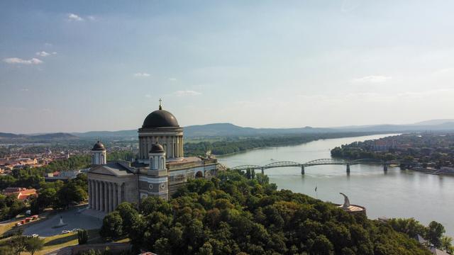 Aerial drone view of Esztergom Basilica with its prominent domed roof overlooking the Danube River. The Mária Valéria Bridge spans the river below, with dense forests framing the historic neoclassical church and surrounding landscape under a blue sky.