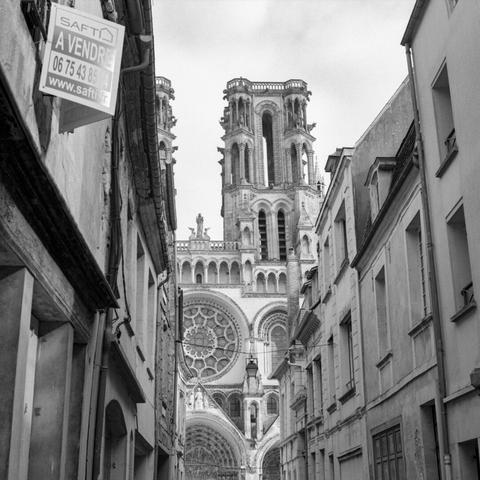 Black and white photo of a narrow street in a historic french city center. The facade and a tower of a gothic cathedral are partially visible at the end of the street.