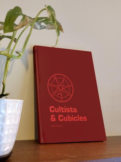 A beautiful, blood red book on a shelf next to a plant. The text on the cover is Cultists & Cubicles, Adam Boman.