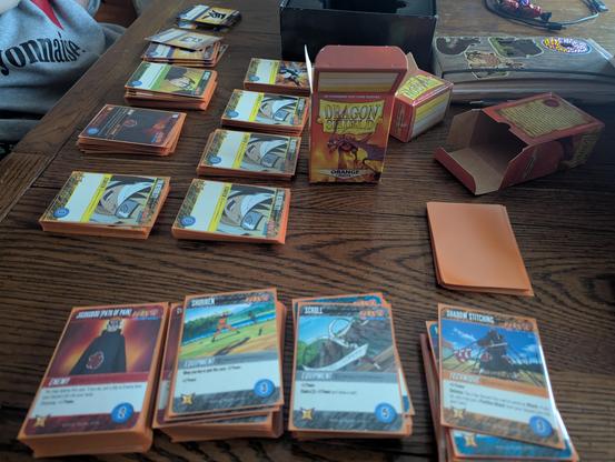 The Cryptozoic Naruto Shippuden Deck Building Game cards laid out on a table, being sleeved with Orange Dragonshield sleeves.