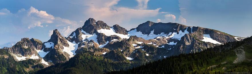 A panorama of a mountain range (the Cowlitz chimneys) with patches of glaciers or snow that has not yet melted. The sky is blue with some clouds.