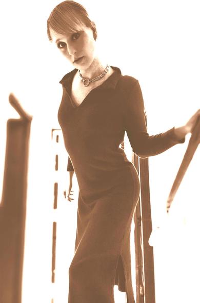 Sepia tone portrait of May wearing a slinky maxi dress. Her hand is resting on a rail and and is looking at the camera.