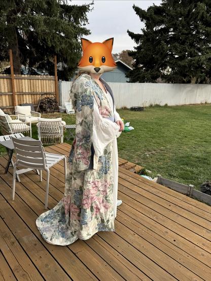 A person wearing a floral-patterned robe stands on a wooden deck. Their face is obscured by a fox emoji. The background features greenery and wooden furniture.