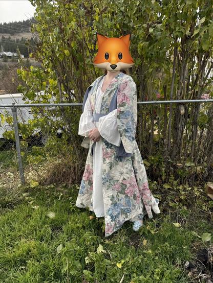 A person stands in a garden, wearing a floral-patterned robe with wide sleeves. Their head is replaced with a cartoon fox emoji. Behind them is a green bush and a fence. The ground is covered with grass and fallen leaves.