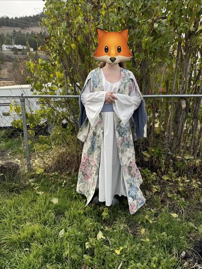 A person is standing in front of a fence, wearing a floral robe over a white dress. Their face is obscured by a fox emoji mask, and they are surrounded by greenery and outdoor scenery.