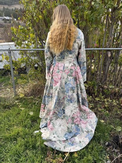 A person with long, wavy hair is standing with their back to the camera, wearing a long floral dress featuring various colors of flowers. They are positioned in a green outdoor area, surrounded by shrubs and a fence.