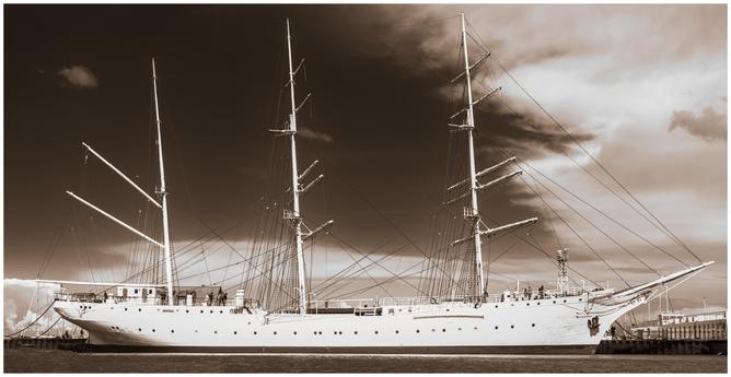 A large, white sailing ship with three tall masts docked at a pier. The rigging is detailed, and multiple ropes extend between the masts and the deck. The ship’s hull features several round portholes. Background includes a cloudy sky and calm water, presented in sepia tone. 
### 
Ein großes, weißes Segelschiff mit drei hohen Masten liegt an einem Pier vor Anker. Die Takelage ist detailreich gestaltet, und zahlreiche Taue spannen sich zwischen den Masten und dem Deck. Der Schiffsrumpf weist mehrere runde Bullaugen auf. Im Hintergrund sind ein bewölkter Himmel und ruhiges Wasser in Sepiatönen dargestellt.