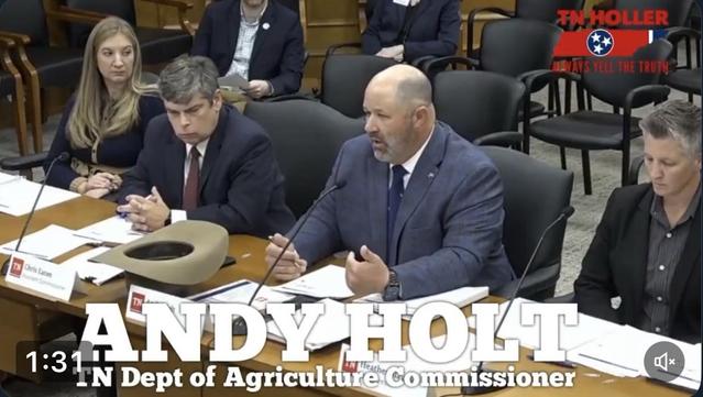 The image shows a meeting setting with several individuals seated around a table, discussing matters related to agriculture. A man in a suit, identified as Andy Holt, is speaking at the table, while others listen attentively. There are nameplates indicating roles.