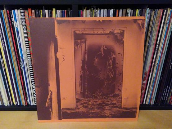 La copertina del vinile appoggiata agli altri vinili. Su una copertina dalla tonalità arancione è raffigurato l'interno di un edificio probabilmente andato a fuoco (pare di vedere, attraverso una porta e intorno a essa, i resti di un incendio).