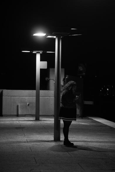 A woman stands under a streetlight at night, illuminated against a dark background. She is looking at her phone, wearing a denim jacket and a skirt. The scene has a moody, monochrome aesthetic with shadows cast on the ground.