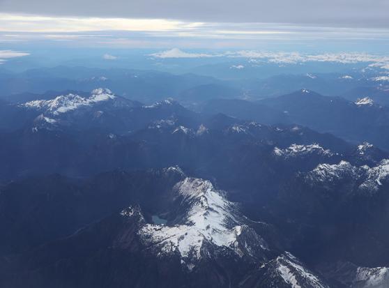 A breathtaking aerial view of a vast mountain range, captured from a high altitude. Snow-capped peaks dominate the foreground, their jagged ridges and valleys casting deep shadows. The mountains stretch into the distance, gradually fading into layers of mist and haze. A small, glacial lake is nestled between the peaks, reflecting the surrounding landscape. The horizon reveals a soft gradient of clouds and sky, with the faint outline of a distant volcano or high summit piercing the cloud layer. The scene is bathed in natural light, creating a serene and expansive atmosphere.