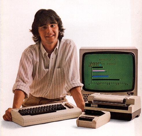 Picture of a young man in a collared shirt with his sleeves rolled up. He is posing with a Commodore VIC-20 computer, along with a datasette tape player, a dot matrix printer and an old CRT monitor.