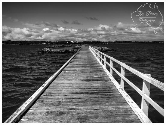 A striking black and white photograph of a wooden jetty extending straight out over a large body of water.

The weathered wooden planks of the jetty, framed by a white railing on the right side, create a strong leading line that draws the eye toward the distant shore and center.

The water is dark and choppy, and the sky is filled with dramatic, bright clouds that add texture and contrast to the scene.

The distant shoreline is dark, providing a strong horizontal line.

The bottom right corner is watermarked "© Kev Peirce."