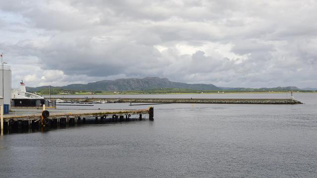 A photo of a pier and breakwater with low rocky mountains in the distance. The sky is filled with clouds.
