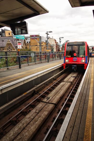 A red Docklands Light Railway (DLR) train, adorned with a remembrance poppy on the front, approaches Tower Gateway station on an elevated track. The train, marked with the number 58, is positioned on the right side of the image, moving towards the platform. The platform is sheltered, with a sign displaying ‘Tower Gateway.’ The urban background includes buildings and a cloudy sky, while the platform is equipped with safety railings and a tactile yellow line for passenger guidance.
