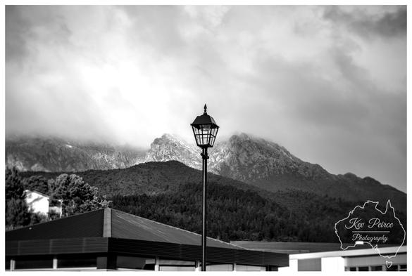 A moody black and white photograph featuring a classic street lamp centered against a dramatic mountain landscape, in Queenstown.

The foreground shows the roofline of a modern building. Behind the light, a forested slope rises to meet rugged, rocky peaks shrouded in low-hanging, textured clouds.

The overall composition creates a strong contrast between the man-made structure and the raw natural environment.