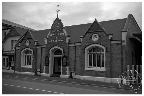 A black and white photograph of a historic brick building with a steeply pitched corrugated iron roof.

The facade features two prominent gabled wings with large arched windows trimmed in white, flanking a central recessed entrance under a smaller gable.

Above the entrance, a stone inscription reads "ZEEHAN SCHOOL OF MINES AND METALLURGY."

A weather vane tops the central gable. Sandwich boards are visible near the entrance, and a road runs along the foreground.