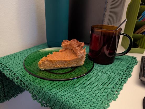 Mesa de escritorio con un tapete verde. Hay una taza de cristal color naranja con el asa en verde y una cuchara. Y también un plato de cristal verde con un trozo de tarta encima. La masa es de hojaldre y el relleno es naranja claro. Parece muy jugoso a juzgar por el aspecto.

De fondo hay algunos elementos como una torre de ordenador, una botella termo verde o un lapicerero con bolígrafos y bolis guardados.