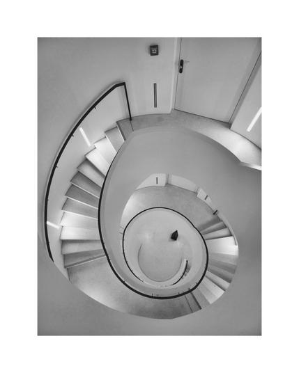 A top-down, black and white photograph of a spiral staircase curving down toward the center. The stairs are solid, light-colored material with darker treads, and they are bordered by a smooth curving wall and a dark handrail. The space is lit, highlighting the edges of the steps. A dark object rests at the bottom center of the spiral. A door and a fire alarm are visible near the top edge.