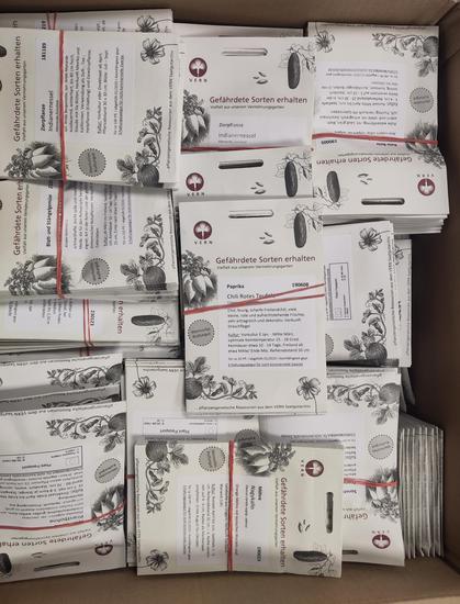 Das Bild zeigt eine Kiste, die dicht gefüllt ist mit zahlreichen, einzeln verpackten Samentüten des Vereins VERN (Verein zur Erhaltung und Rekultivierung von Nutzpflanzen). Die Tüten sind überwiegend weiß, rot und schwarz bedruckt und enthalten Informationen über die jeweiligen Pflanzensorten sowie handgezeichnete Abbildungen von Pflanzen.