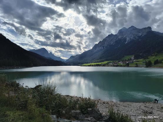 Blick auf einen See in der Schweiz. Im Hintergrund links und rechts die Berge der Alpen. Ein bewölkter Himmel. Die Sonnenstrahlen scheinen durch die Wolken. Der Himmel spiegelt sich im See.
