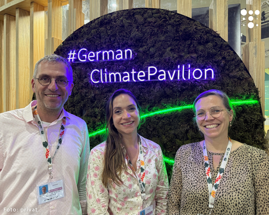 Drei Personen vor einer moosbedeckten Wand mit leuchtendem Schriftzug '#GermanClimatePavilion' und grünem Lichtstreifen.