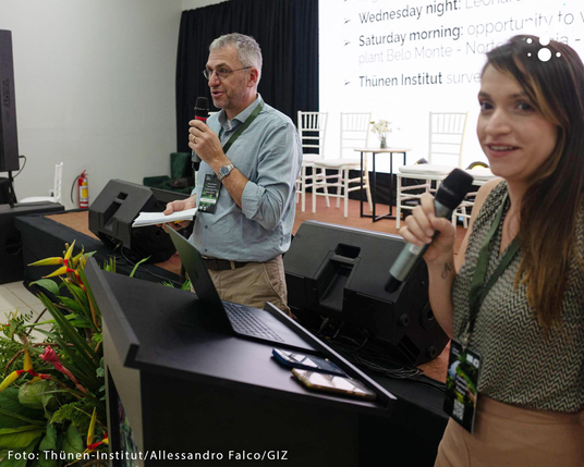 Zwei Personen mit Mikrofonen stehen an einem Rednerpult vor einer Leinwand mit Text, im Hintergrund weiße Stühle und eine an die Wand projizierte Folie einer Präsentation.