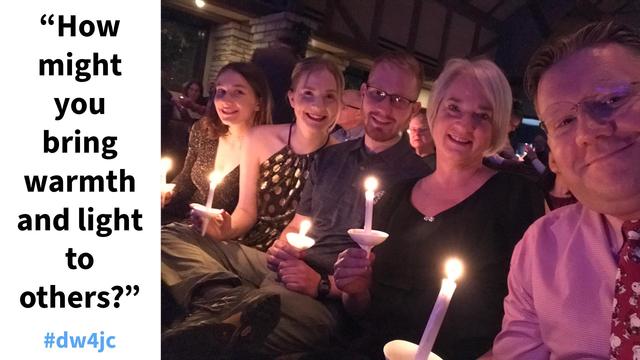 A smiling family of five sits together in a warmly lit church sanctuary during a candlelight service, each holding a glowing candle with white paper drip protectors. The soft candlelight illuminates their faces as they sit close together, surrounded by other attendees in the background. On the left side of the image, white text reads: “How might you bring warmth and light to others?” followed by the hashtag #dw4jc in blue.