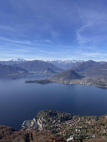 Una vista panoramica di un lago sereno circondato da montagne, con un cielo azzurro e alcune cime innevate in lontananza. Una piccola città con case è visibile lungo il bordo del lago, insieme alle barche sull'acqua.