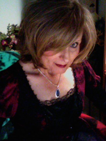 In a dark, moody image, an older woman with her hair down and falling forward, gazes at the camera with only one eye visible, her expression caught between seriousness and playfulness. She's wearing a blood red and black, Victorian style dress and a necklace with a deep purple pendant.