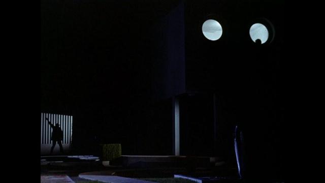 Dark, nighttime. We see the eyes of villa Arpel (windows) lit in the back of the still.
On the left Monsieur Hulot is trying to get in the modern house. The rest of the picture is dark.