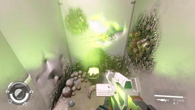 A damaged bathroom stall in an office building. The walls are dented and there are sections covered in an organic compound and what appears to be egg sacs. The toilet lid looks like it exploded and there is a green light being emitted. On the wall over the toilet it says "Don't eat the xeno tacos." with a skull drawn next to it. A rifle can be seen in the foreground as if being carried by the viewer.