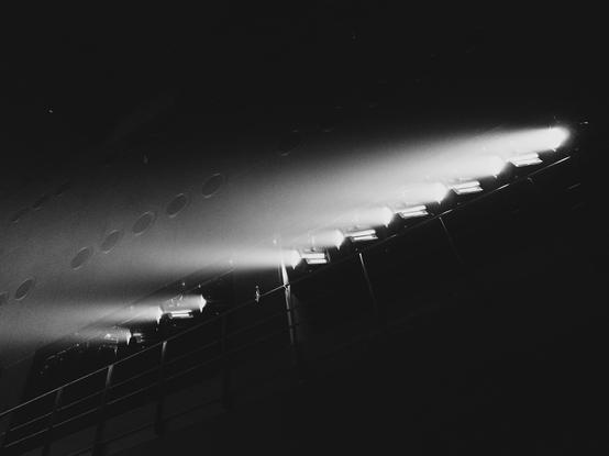 Beam of light cutting through darkness from a row of aligned spotlights, angled downwards, creating sharp contrast in a dark setting. Series of circular holes visible in the background.