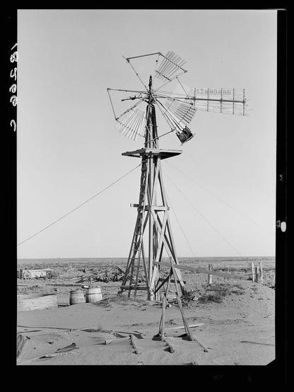 The image depicts a tall windmill standing in an arid landscape. The structure is composed of wooden beams and metal blades, with one blade folded downwards as if it has stopped spinning or is broken. Surrounding the base of the windmill are several barrels, some overturned on their sides, suggesting neglect or disuse. In the background, a desolate field stretches out to meet an overcast sky, indicative of harsh weather conditions typical in rural farming regions during dry seasons. The absence of human presence and structures like wooden fences further emphasizes abandonment.