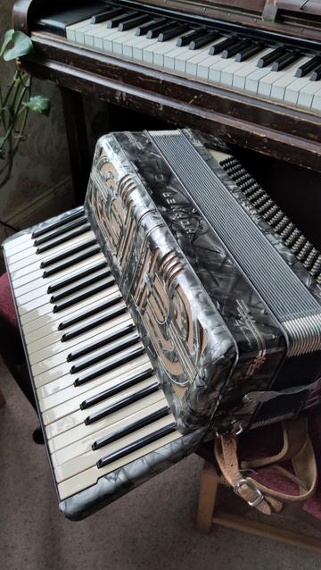 A large piano accordian sitting in front of a piano. It is in shades of perlescent grey and sort of art deco style. One side has piano keys, the other has a very prolific bank of black buttons.