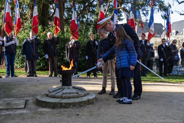 Des enfants rallument la flamme du souvenir, avec le délégué général du Souvenir Français de Loir-et-Cher, le général Beyer, sous les arbres de la place de la République, devant le monument aux morts hors champ, avec les porte-drapeaux des associations patriotiques en arrière-plan.