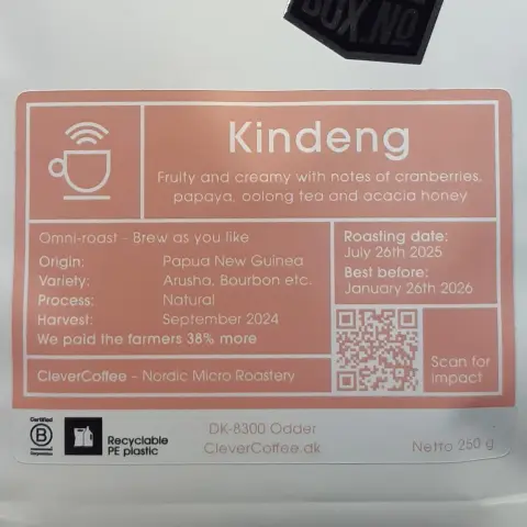 The image shows a coffee package labeled "Kindeng." The background is mainly white with some red accents. The top features a simple illustration of a coffee cup. Below it, the text reads "Fruity and creamy with notes of cranberries, papaya, oolong tea, and acacia honey." The package details include information about the origins, mentioning "Omni-roast - Brew as you like," and specifying "Origin: Papua New Guinea," with the variety as "Arusha, Bourbon etc." The processing date is listed as "September 2024," and there's a note about the roast date being "July 26th 2025." A QR code is present for scanning. The bottom section states "CleverCoffee - Nordic Micro Roastery," and it shows the address "DK-8300 Odder" along with a net weight of "250 g." Additionally, there’s a note about the packaging being recyclable PE plastic.