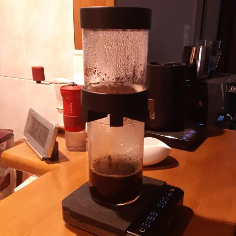 The image shows a coffee brewing device set on top of a glass, which is resting on digital scales. The brewing device has a transparent upper chamber filled with coffee grounds, and a glass partially filled with brewed coffee. The scales display a digital readout, showing the weight and time. In the background, there are various kitchen items like a red manual coffee grinder and a goose-neck kettle.