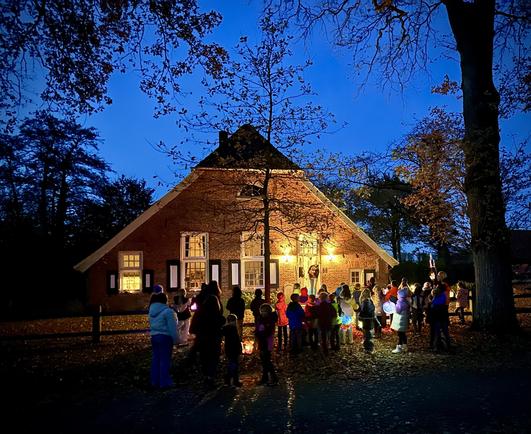 Das Bild zeigt ein braunes Gebäude mit einem steilen Dach, das in einem dunklen Wald steht. Eine große Kindergruppe steht vor dem Gebäude und blickt darauf. Einige Personen halten Lichtquellen, wie Laternen oder Taschenlampen. Das Gebäude hat mehrere Fenster und eine Tür in der Mitte. Die Umgebung ist dunkel, aber das Gebäude ist durch die Lichtquellen und die Beleuchtung im Inneren sichtbar.