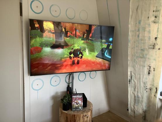 A TV in the corner with a video game playing on a computer on top of a tree trunk. Brightly colored nature scene.