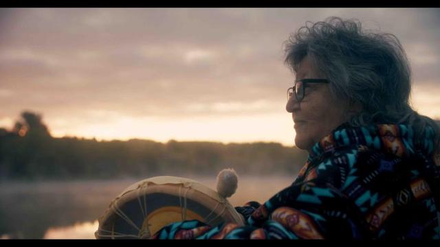 Photo of an elderly woman sitting near the water at sunset and holding a drum, smiling at something in the distance.
