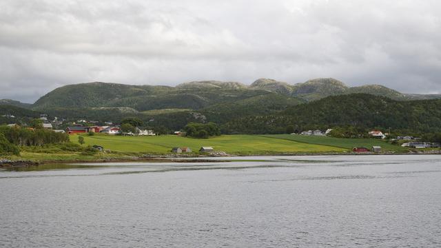 A photo of farm fields on a shore leading into forests with low mountains in the distance. The sky is cloudy.