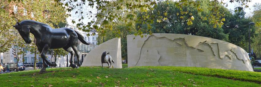 A photo of the back of Animals in War memorial in London, England. A bronze sculpture of a horse and a dog walk away from the gap in the memorial. The sculpture of the dog is looking back at it.