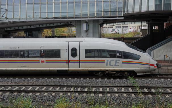 ICE 3 mit Regenbogenstreifen von der Seite. Dahinter die Beton- und Glaskonstruktion des Bahnhofs