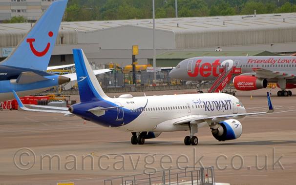 Side view of a twin engined jet airliner taxiing from left to right at a busy airport.
The plane is largely white, with large, dark blue "Kuwait" titles on the forward fuselage just aft of the forward door, and a small Kuwaiti flag just forward of the door.
A large, blue, stylised bird covers the rear of the tail and lower rear fuselage, with the black registration "9K-AKM" on the upper rear fuselage, over the birds head.
A smaller, white bird is also on the tail.
The white engine pods under the wing have a diagonal blue band around the rear, and a dark blue flying bird at the front.#
Grey concrete apron fills the foreground, with the tail of a blue jet airliner on the left of the background, and the nose and forward fuselage of a grey and red jet airliner on the right, both parked facing grey cargo sheds, with trees behind that.