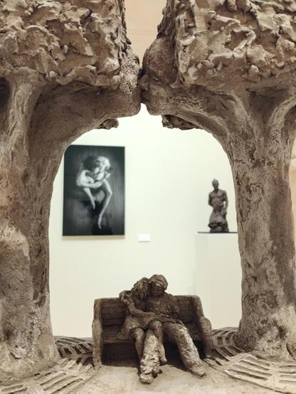 Au premier plan, une sculpture représentant un couple sur un ban public entouré de 2 arbres qui s'embrassent au dessus du couple. En arrière plan, une photo en noir et blanc représentant un homme jouant du ukulélé et une sculpture de buste d'homme en terre noire.