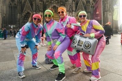 Vier Frauen in quietschbunten Overalls, bunte brillen, eine trägt aufgeblasenes Luftkissen in Form eines Ghettoblasters.die Gruppe soll vielleicht Hip-Hoper darstellen.