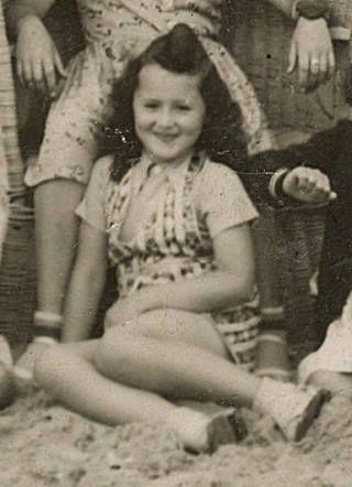 A young smiling girl sitting on sand. Some people visible in the background.